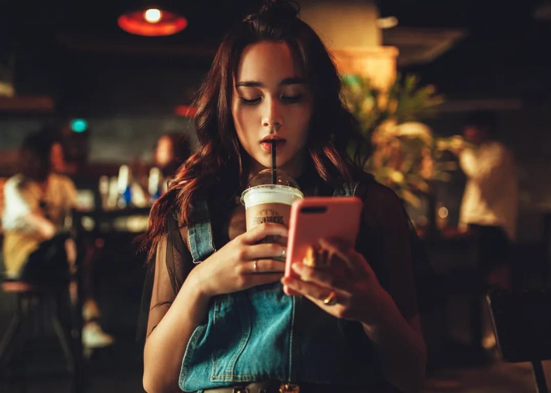 A woman looking at her phone with a drink in a warmly lit cafe